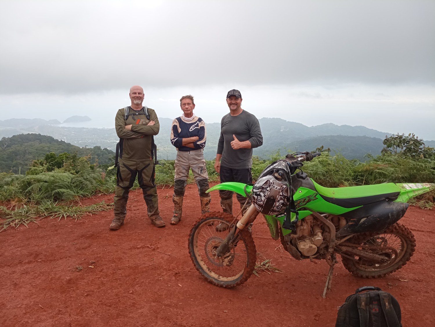 Three riders with a dirt bike on a red-dirt mountain trail overlooking the Costa Rican coastline